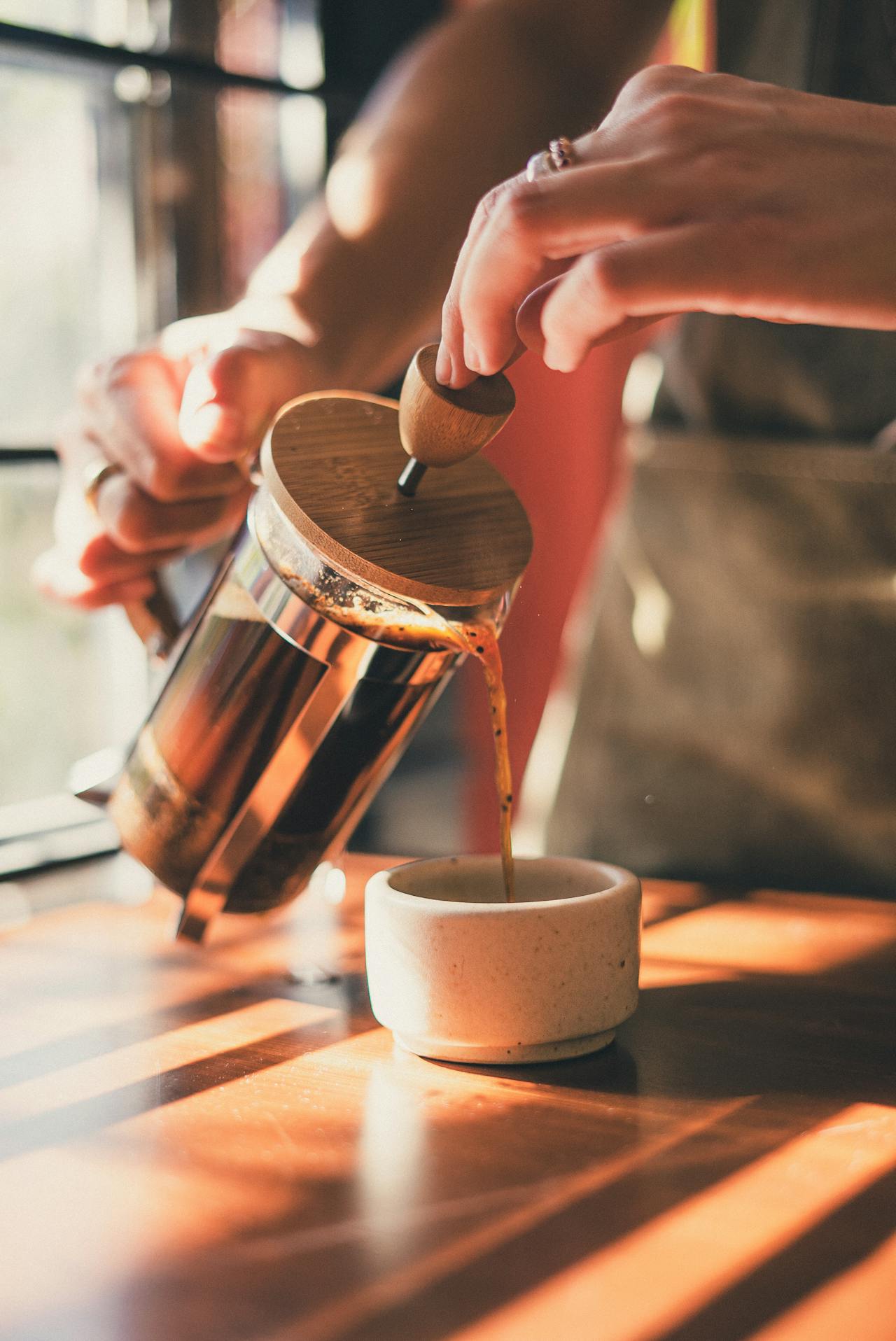 Hands holding French press at 45 degree angle, pouring coffee into small stone cup with light shining through a window in the background.